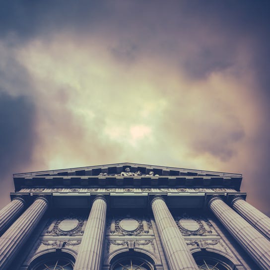 Dramatic upward view of a classical courthouse with tall stone columns and detailed carvings, set against a cloudy, moody sky.
